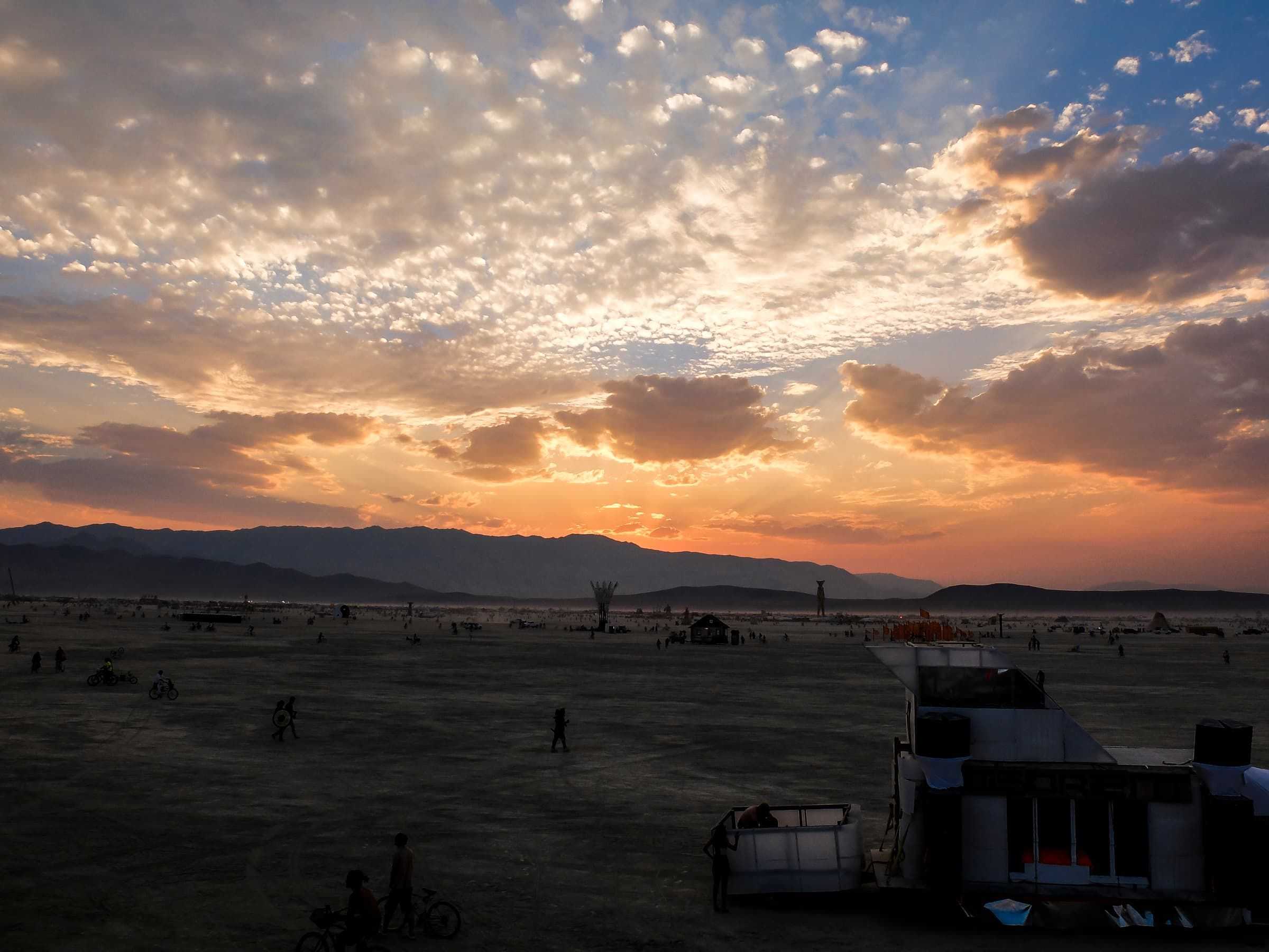 Open alkali flats of the Black Rock Desert at sunset, with a low sun and a figure walking toward a distant camp.